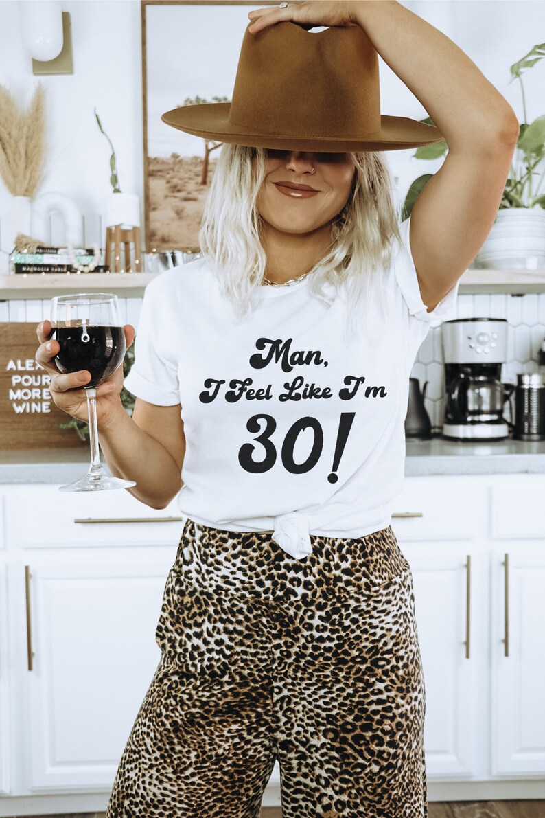 May include: A white t-shirt with the text "Man, I feel like I'm 30!" in a retro font. The person wearing the shirt is holding a glass of red wine and wearing a brown fedora and leopard print pants.