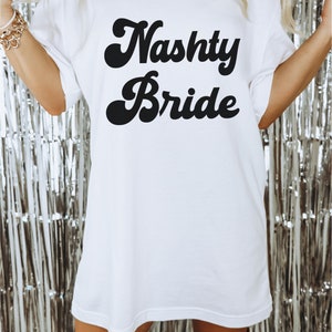Pu&ograve; includere: Maglietta bianca oversize con testo nero che recita "Nasty Bride" in un font retr&ograve;.