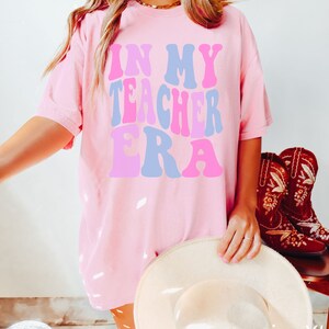 May include: Pink oversized t-shirt with the text "In My Teacher Era" in a retro font with pink, blue, and purple colours. The shirt is shown on a model wearing brown leggings and a white hat. The text "size XL" and "BLOSSOM" are visible in the bottom right corner of the image.