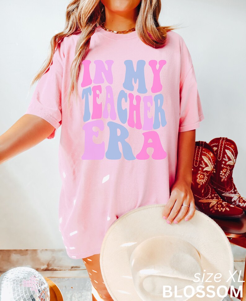 May include: Pink oversized t-shirt with the text "In My Teacher Era" in a retro font with pink, blue, and purple colours. The shirt is shown on a model wearing brown leggings and a white hat. The text "size XL" and "BLOSSOM" are visible in the bottom right corner of the image.