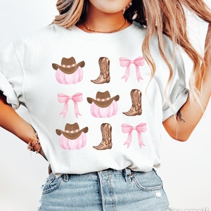 May include: White t-shirt with a repeating pattern of cowboy hats, boots, and bows. The cowboy hats have star accents, and the boots are brown. The bows are pink. The text "size small WHITE" is at the bottom right.