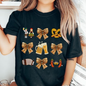 Op de afbeelding: Zwart T-shirt met een feestelijk Duits thema. Het ontwerp bevat een stel in traditionele kleding, pretzels, bierglazen, een strik, een accordeon en twee worstjes die bierglazen vasthouden.