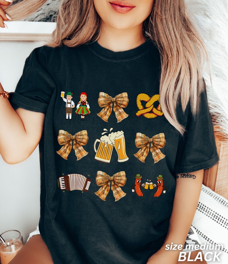 Op de afbeelding: Zwart T-shirt met een feestelijk Duits thema. Het ontwerp bevat een stel in traditionele kleding, pretzels, bierglazen, een strik, een accordeon en twee worstjes die bierglazen vasthouden.
