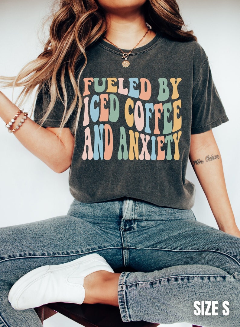 Peut inclure: T-shirt gris fonc&eacute; avec le texte "FUELED BY ICED COFFEE AND ANXIETY" dans des lettres color&eacute;es de style r&eacute;tro. Le t-shirt est associ&eacute; &agrave; un jean bleu et des baskets blanches. La taille est S.