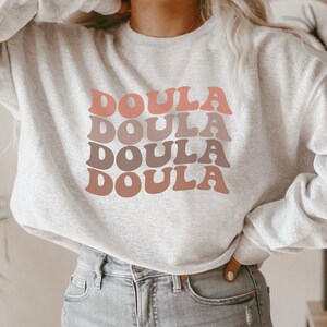 Op de afbeelding: Een grijs sweatshirt met het woord "Doula" vier keer herhaald in een retro-lettertype, waarbij elk woord in een andere tint roze en bruin is.