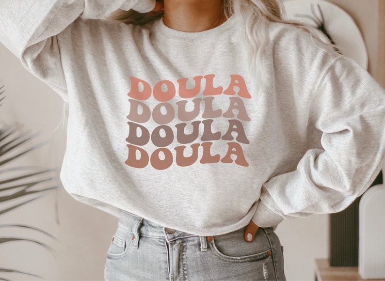Op de afbeelding: Een grijs sweatshirt met het woord "Doula" vier keer herhaald in een retro-lettertype, waarbij elk woord in een andere tint roze en bruin is.