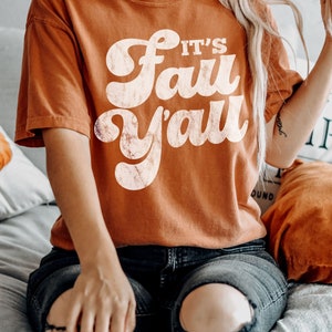 May include: A woman wearing a burnt orange t-shirt with the text "It's Fall Y'all" in white retro font. She is wearing ripped jeans and sitting on a bed with a white blanket.