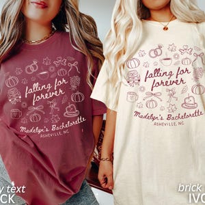 May include: Two t-shirts, one brick red and one ivory, with the text "falling for forever" and "Madelyn's Bachelorette Asheville, NC". The shirts have a variety of small graphic illustrations.