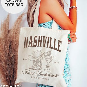 May include: Natural canvas tote bag with "Nashville" text and a cowboy hat, boots, and cocktail illustration. Features "Aria's Bachelorette" and "Let's Go Girls" text. Bag dimensions are 15" x 16" and made of 100% cotton.