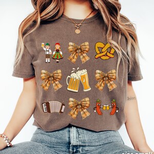 Op de afbeelding: Een bruin T-shirt met een feestelijk Oktoberfest-thema. Het ontwerp bevat een stel in traditionele Duitse kleding, pretzels, bierkruiken, accordeons en strikken met ruitjespatronen.