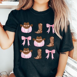 May include: Black t-shirt featuring a repeating pattern of pink pumpkins with brown cowboy hats, brown cowboy boots, and pink bows. The shirt has the text "size medium BLACK" at the bottom.