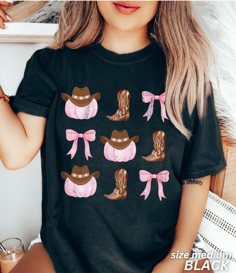 May include: Black t-shirt featuring a repeating pattern of pink pumpkins with brown cowboy hats, brown cowboy boots, and pink bows. The shirt has the text "size medium BLACK" at the bottom.