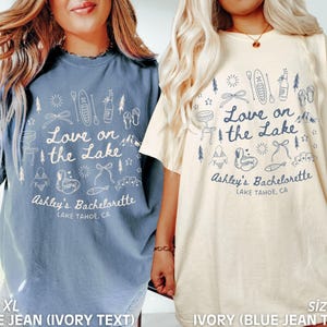 May include: Two XL t-shirts, one blue jean with ivory text and one ivory with blue jean text. Each shirt has a graphic that says "Love on the Lake" with lake-themed illustrations and "Ashley's Bachelorette, Lake Tahoe, CA."