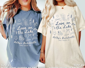 Op de afbeelding: Twee XL-T-shirts, één blauwe jeans met ivoorkleurige tekst en één ivoorkleurige met blauwe jeans-tekst. Elk shirt heeft een afbeelding met de tekst "Love on the Lake" met illustraties met een meer-thema en "Ashley's Bachelorette, Lake Tahoe, CA."