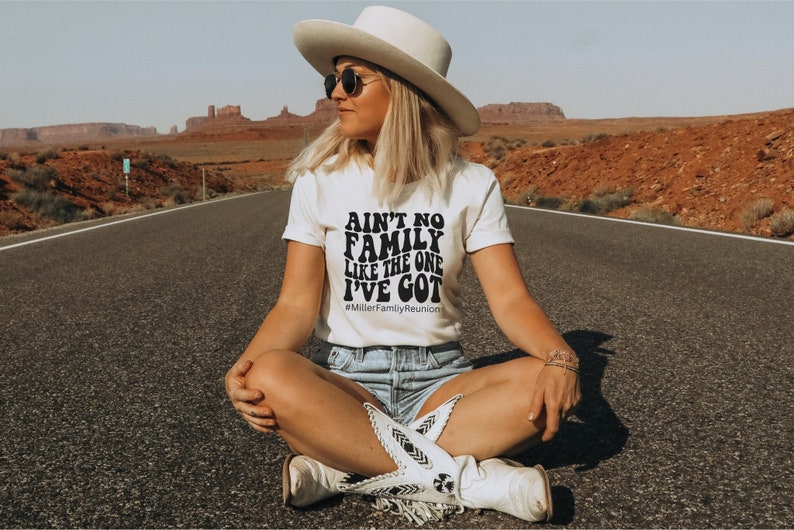 May include: A white t-shirt with black text that reads "Ain't no family like the one I've got" and the hashtag "#MillerFamilyReunion". The person wearing the shirt is sitting on a paved road in a desert landscape.