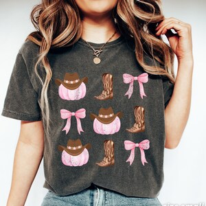 May include: Dark gray t-shirt featuring a repeating pattern of pink pumpkins with brown cowboy hats, brown cowboy boots, and pink bows. The text "size small PEPPER" is printed in white at the bottom right.