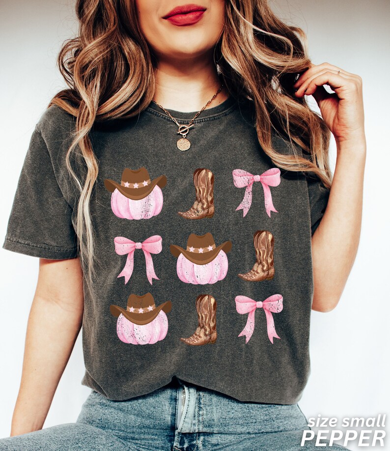 May include: Dark gray t-shirt featuring a repeating pattern of pink pumpkins with brown cowboy hats, brown cowboy boots, and pink bows. The text "size small PEPPER" is printed in white at the bottom right.