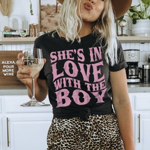 Puede incluir: Una mujer con una camiseta negra con el texto "She's in love with the boy" en letras rosas. Lleva un sombrero marr&oacute;n y pantalones de estampado de leopardo. Est&aacute; sosteniendo una copa de vino blanco.