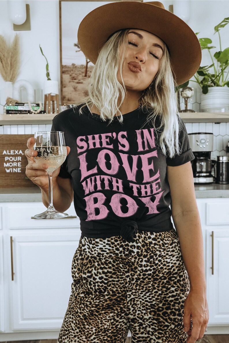 Puede incluir: Una mujer con una camiseta negra con el texto "She's in love with the boy" en letras rosas. Lleva un sombrero marr&oacute;n y pantalones de estampado de leopardo. Est&aacute; sosteniendo una copa de vino blanco.