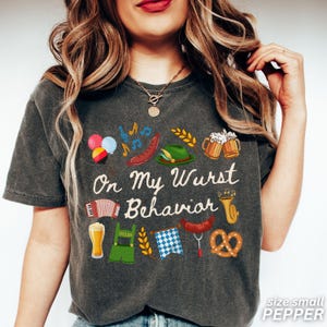 May include: Dark gray t-shirt with the phrase "On My Wurst Behavior" and various Oktoberfest-themed illustrations. The design includes beer steins, sausages, pretzels, and an accordion. The shirt is size small.