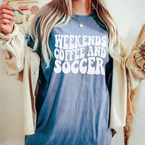 May include: A blue jean oversized t-shirt with the text "Weekends Coffee and Soccer" in white, retro-style font. The shirt is shown on a model and the text "Size XL - Blue Jean" is visible at the bottom of the image.