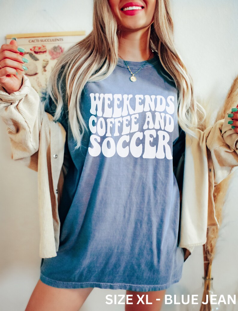 May include: A blue jean oversized t-shirt with the text "Weekends Coffee and Soccer" in white, retro-style font. The shirt is shown on a model and the text "Size XL - Blue Jean" is visible at the bottom of the image.