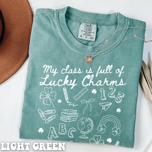 May include: Light green t-shirt with the text "My class is full of Lucky Charms" and white illustrations of school supplies, shamrocks, and the numbers 1, 2, and 3.