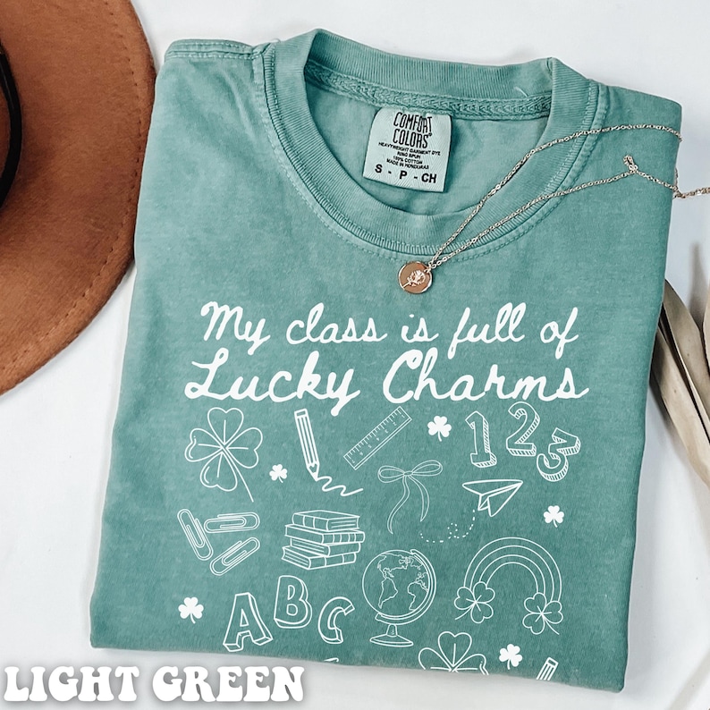 May include: Light green t-shirt with the text "My class is full of Lucky Charms" and white illustrations of school supplies, shamrocks, and the numbers 1, 2, and 3.