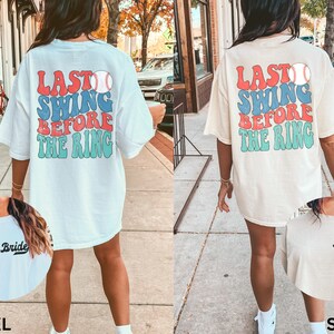 May include: Two white oversized t-shirts with a baseball graphic and the text "Last Swing Before The Ring". One shirt is white and the other is ivory. The shirts have the words "Bride" and "Babe" printed on the front.