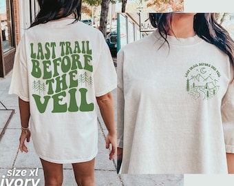 May include: Ivory oversized t-shirt with a green graphic on the back that says "Last Trail Before The Veil" and a smaller green graphic on the front with a mountain scene and the text "Last Trail Before The Veil".