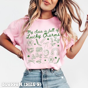 May include: Pink t-shirt with a green graphic that says "My class is full of Lucky Charms" with school-themed icons like a pencil, ruler, books, globe, and a rainbow.