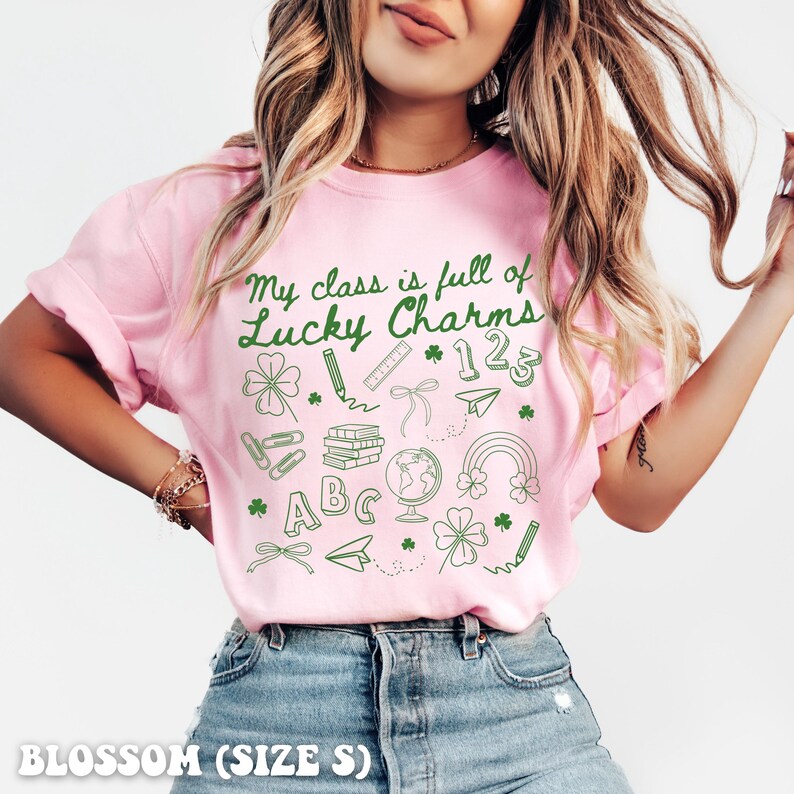 May include: Pink t-shirt with a green graphic that says "My class is full of Lucky Charms" with school-themed icons like a pencil, ruler, books, globe, and a rainbow.