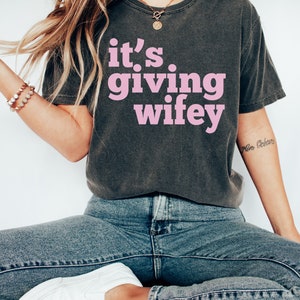May include: A dark gray t-shirt with the text "it's giving wifey" in pink lettering.