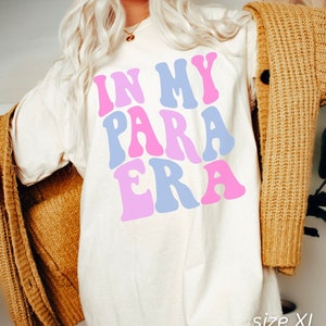 May include: A white oversized t-shirt with the text "IN MY PARA ERA" in pink, blue, and light pink letters. The shirt is shown on a model wearing a brown cardigan. The text "size XL IVORY" is visible at the bottom of the image.
