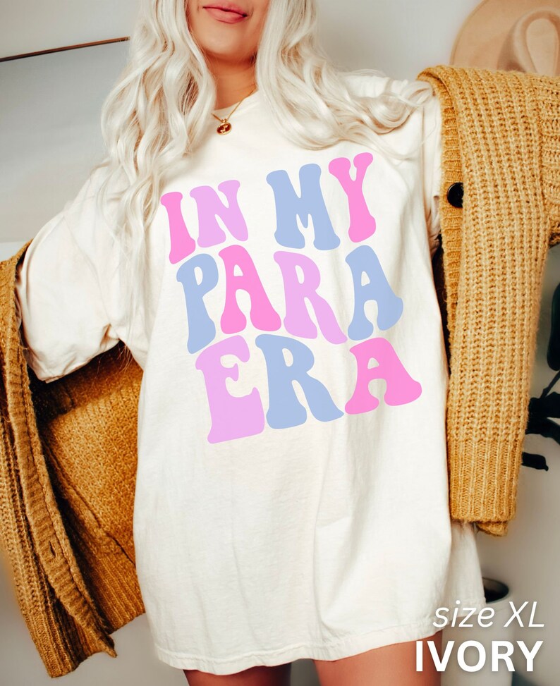 May include: A white oversized t-shirt with the text "IN MY PARA ERA" in pink, blue, and light pink letters. The shirt is shown on a model wearing a brown cardigan. The text "size XL IVORY" is visible at the bottom of the image.