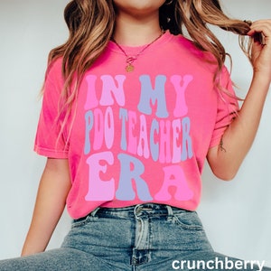 Peut inclure: Un t-shirt rose avec le texte "In My P.D.O Teacher Era" dans une police r&eacute;tro avec des couleurs rose, violet et bleu.