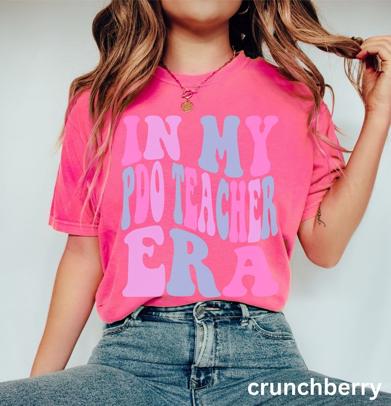 Peut inclure: Un t-shirt rose avec le texte "In My P.D.O Teacher Era" dans une police r&eacute;tro avec des couleurs rose, violet et bleu.