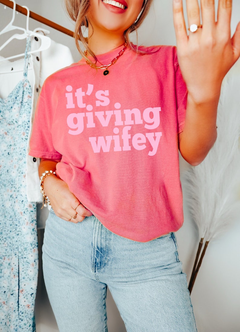 May include: A pink t-shirt with the text "it's giving wifey" in pink lettering.