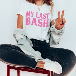 K&ouml;nnte beinhalten: Wei&szlig;es T-Shirt mit dem Text "MY LAST BASH" in Pink. Das Shirt wird mit dunklen Jeans, einer hellen Jeansjacke und wei&szlig;en Schuhen getragen. Die Person sitzt auf einem Holzschemel.