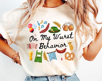 Op de afbeelding: Crèmekleurig T-shirt met de tekst "On My Wurst Behavior" en diverse Duitse illustraties. Het ontwerp bevat worsten, bierpullen, een pretzel, een accordeon en een traditionele hoed.