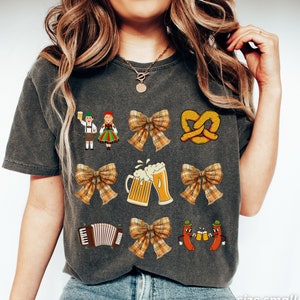 Op de afbeelding: Een grijs T-shirt met verschillende kleurrijke illustraties, waaronder een pretzel, bierglazen, een strik, een accordeon en twee figuren in traditionele Duitse kleding. De tekst "PEPPER" staat gedrukt in de rechterbenedenhoek van het shirt.