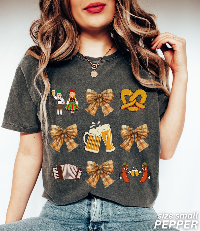 Op de afbeelding: Een grijs T-shirt met verschillende kleurrijke illustraties, waaronder een pretzel, bierglazen, een strik, een accordeon en twee figuren in traditionele Duitse kleding. De tekst "PEPPER" staat gedrukt in de rechterbenedenhoek van het shirt.