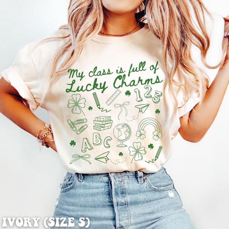 May include: Ivory t-shirt with the text "My class is full of Lucky Charms" in green lettering. The shirt features a variety of green shamrock and school-themed illustrations.