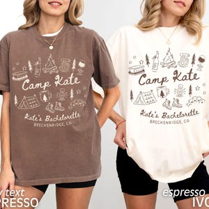 May include: Two short-sleeved t-shirts, one brown and one ivory, each with a white graphic. The graphic includes the text "Camp Kate" and "Kate's Bachelorette, Breckenridge, CO" along with camping-themed illustrations like a tent, campfire, and hiking boots.
