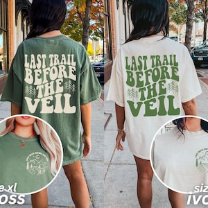 Puede incluir: Dos camisetas de gran tamaño con el texto "Last Trail Before The Veil" en una fuente retro. Las camisetas son de color verde musgo y marfil con un pequeño gráfico en la parte delantera de cada camiseta. El texto en la parte posterior de las camisetas es de color crema.
