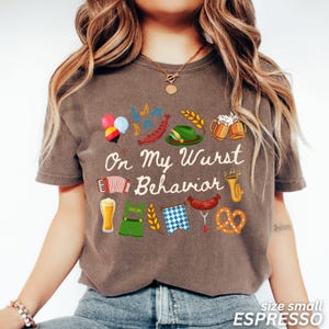May include: Espresso-colored t-shirt with the phrase "On My Wurst Behavior" and festive German-themed graphics. The design includes beer steins, pretzels, sausages, and an accordion. The shirt is size small.