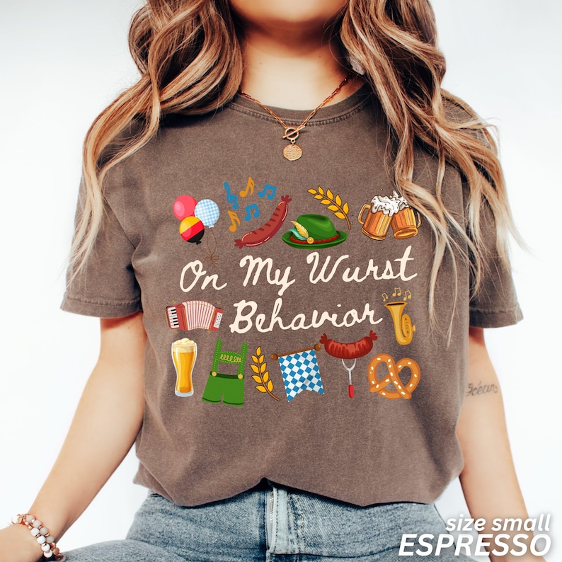 May include: Espresso-colored t-shirt with the phrase "On My Wurst Behavior" and festive German-themed graphics. The design includes beer steins, pretzels, sausages, and an accordion. The shirt is size small.