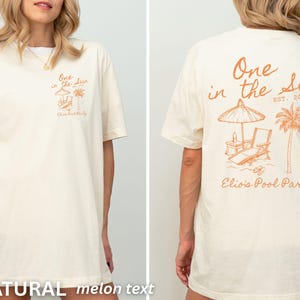 Op de afbeelding: Cr&egrave;mekleurig T-shirt met een oranje strandthema. De voorkant heeft tekst en een kleine illustratie, de achterkant een groter ontwerp met een parasol, stoel en palmboom. Tekst luidt "One in the Sun" en "Elio's Pool Party".