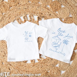 Op de afbeelding: Twee witte T-shirts met korte mouwen, met een blauwe afbeelding op de voor- en achterkant. Het ontwerp bevat de tekst "One in the Sun" met een strandsc&egrave;ne en "Elisa's Pool Party". De shirts worden gepresenteerd op een geweven mat.