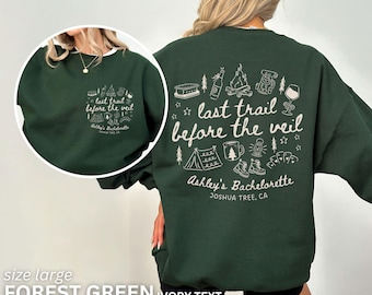 May include: A forest green sweatshirt with ivory text. The front features a small graphic with the text "last trail before the veil" and "Ashley's Bachelorette, Joshua Tree, CA". The back has a larger graphic with camping-themed illustrations.